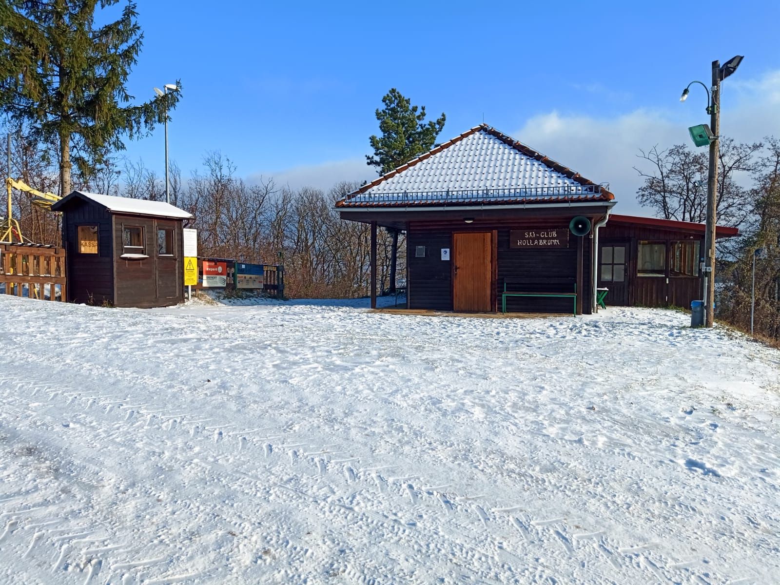 Schneebedeckte Schiclub-Hütte mit Holzhäuschen und Bäumen im Hintergrund.