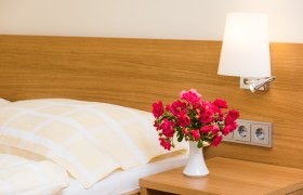 Ein G&auml;stezimmer mit einem Bett, einer Nachttischlampe und einem Blumenstrau&szlig; in einer Vase auf dem Nachttisch.