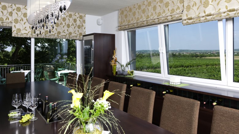 Ein Verkostungsraum mit einem großen Tisch, Weingläsern und Flaschen. Große Fenster bieten Blick auf Weinberge. Elegante Dekoration und Beleuchtung.