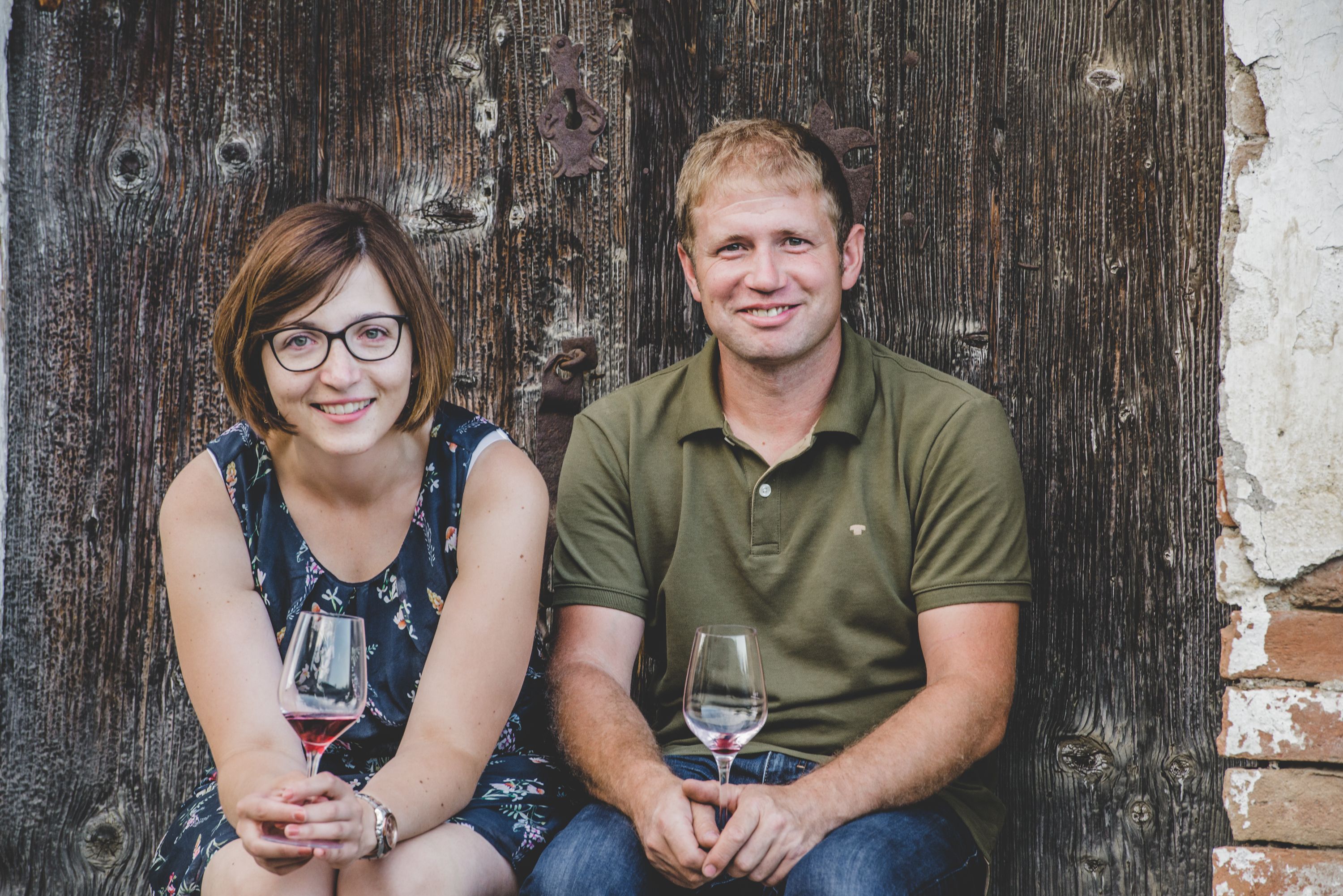 Ein Mann und eine Frau sitzen vor einer Holzwand und halten Weingläser in den Händen.