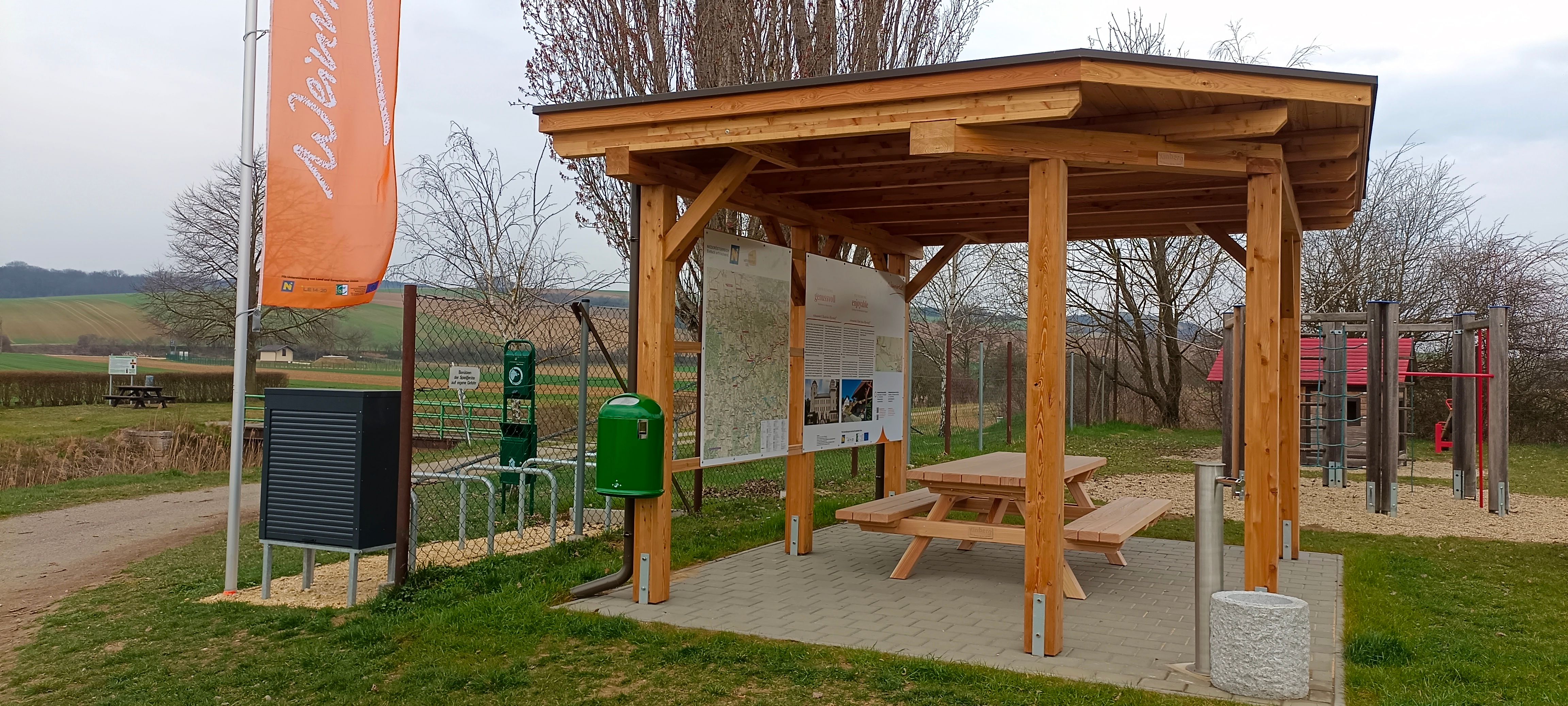 Ein Rastplatz mit Holzdach, Tisch und Bank, umgeben von Informationstafeln und einem Spielplatz im Hintergrund.