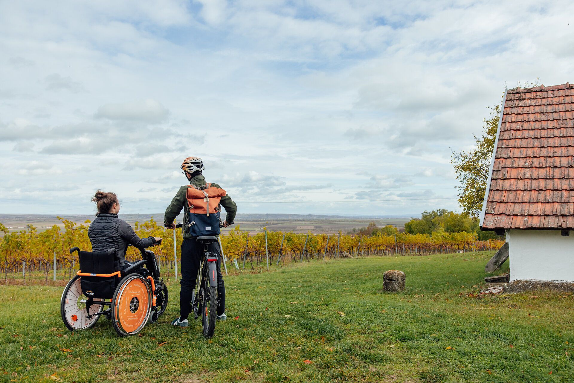 Eine Frau im E-Rollstuhl mit Zusatzrad und ein Mann auf einem Fahrrad genießen den Ausblick über die mit herbstlichen Weingärten durchzogene Weinviertler Landschaft. Am Bildrand sieht man im Anschnitt ein Presshaus.