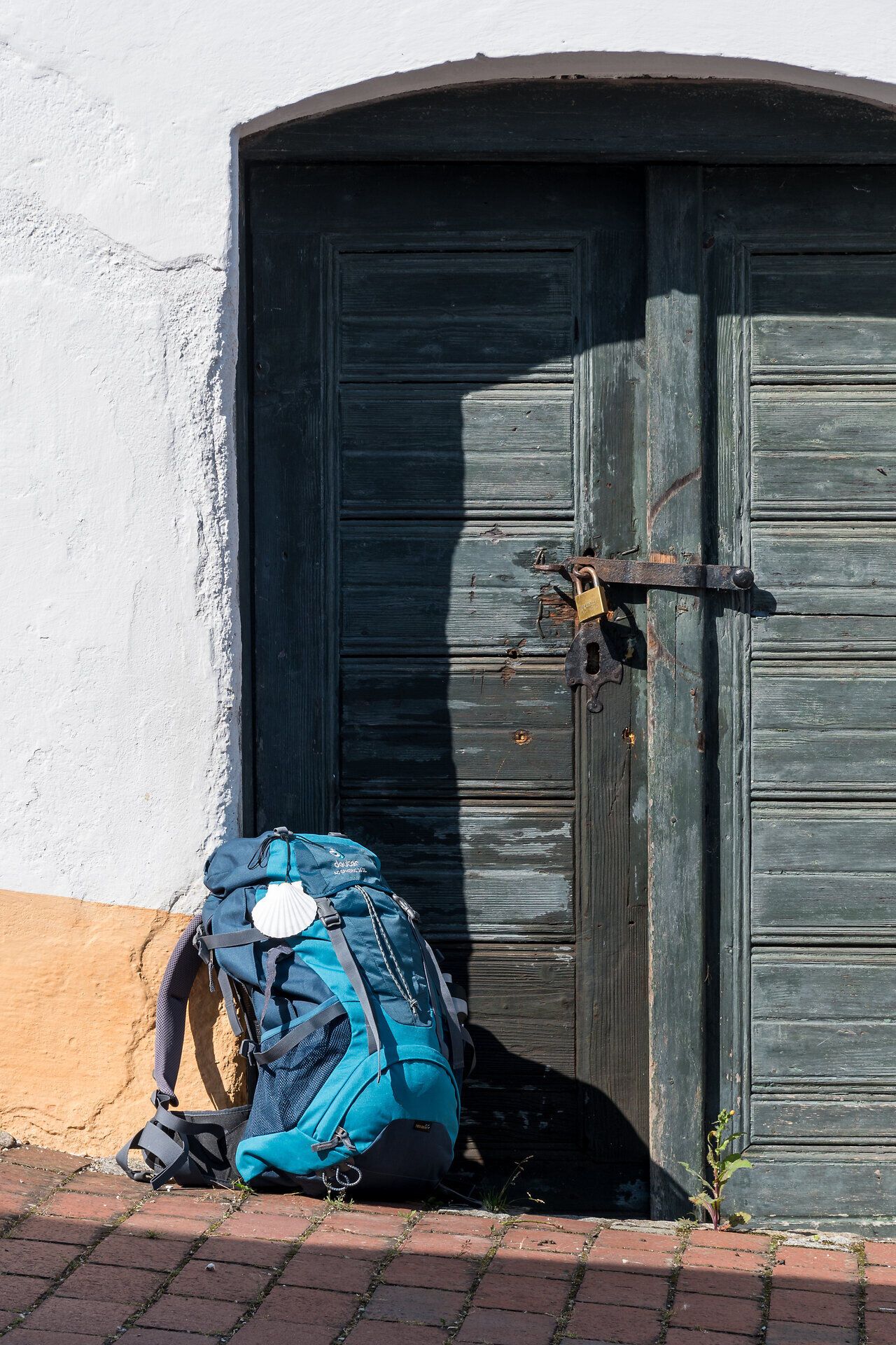 Eine rustikale Kellertür, umgeben von der warmen Sonne, strahlt eine einladende Atmosphäre aus. Neben der Tür lehnt ein Rucksack, der auf Abenteuer und Erkundungen in der Umgebung hinweist. Die sanften Farben der Wand und die Schatten der Tür schaffen eine harmonische Kulisse für Reisende.