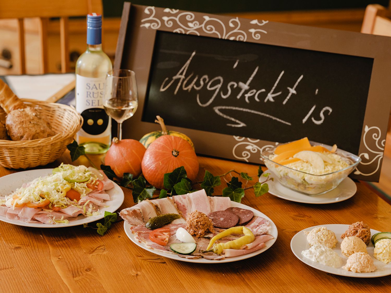 Ein gedeckter Tisch mit kalten Platten, Brot, Wein und einem Schild mit der Aufschrift 'Ausg'steckt is'.