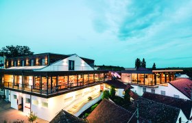 Moderne Weingut-Architektur bei Dämmerung mit beleuchteten Innenräumen und umliegenden Gebäuden.