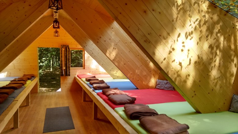 Innenansicht eines Holzhütten-Schlafsaals mit Matratzen und Decken.