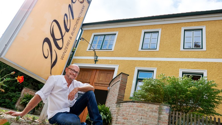 A man sits smiling in front of a yellow building with a sign that reads '20ers Winzerei & Buschenschank (typical tavern)'.