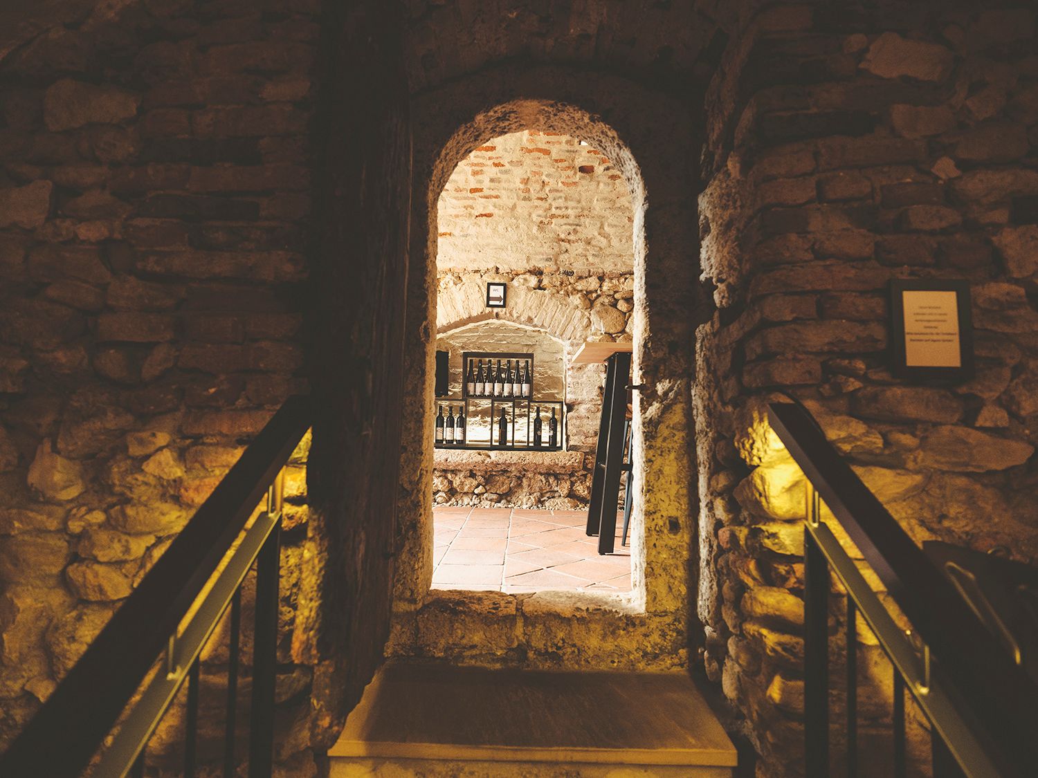 Steinmauer mit Durchgang zu einem Raum mit Flaschen auf einem Regal.