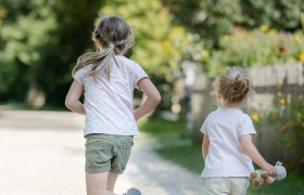 Zwei Kinder laufen auf einem Kiesweg in einem Park, umgeben von Bäumen und Blumen.