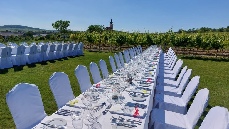 Lange, festlich gedeckte Tafel im Freien mit weißen Stühlen, umgeben von Weinbergen und einem Kirchturm im Hintergrund.