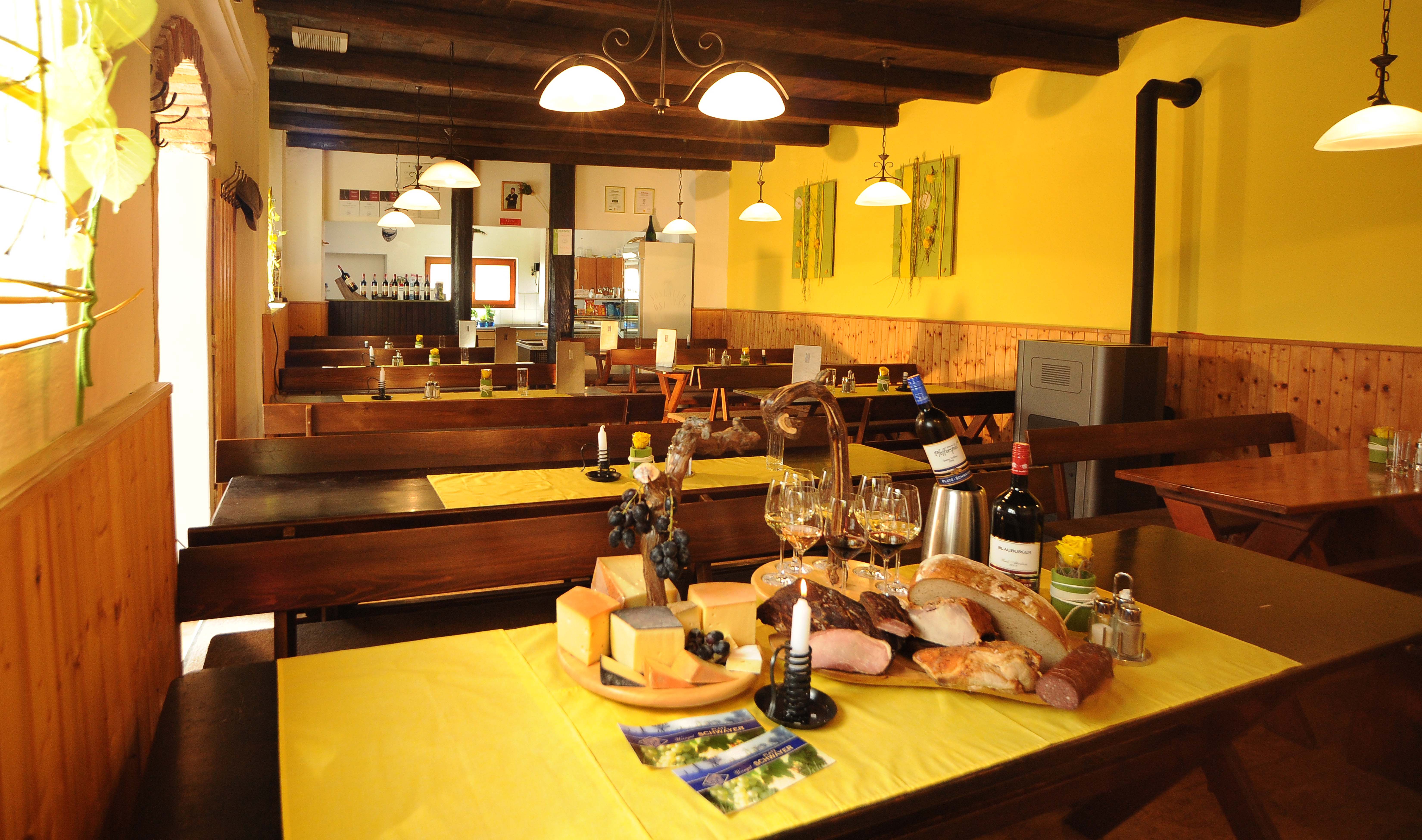 Innenansicht eines gemütlichen Heurigen mit Holztischen, gelben Tischdecken und einer Auswahl an Käse, Brot und Wein auf einem Tisch.