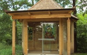 Kleine Holzkapelle mit Glasfront und Kreuz auf dem Dach, umgeben von Bäumen.