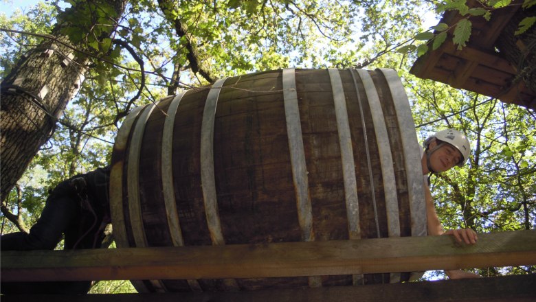 Person mit Helm kriecht durch ein großes Holzfass in einem Kletterpark.