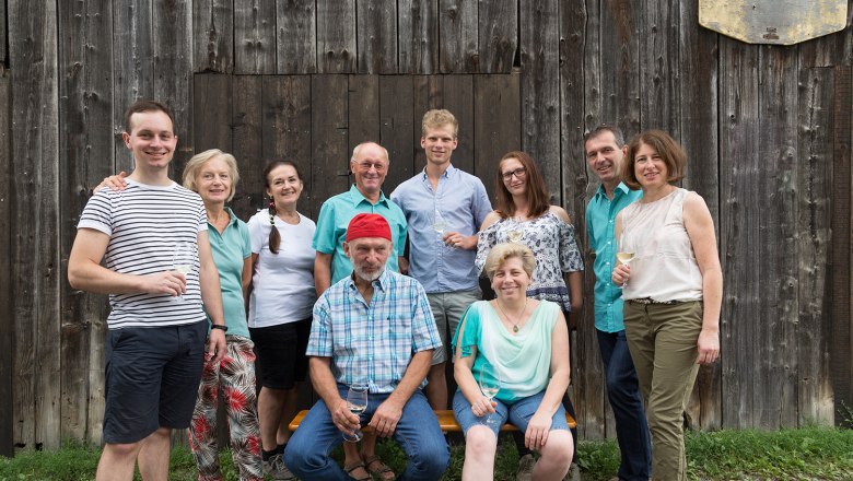 Eine Gruppe von zehn Personen posiert l&auml;chelnd vor einer Holzwand, einige halten Weingl&auml;ser.