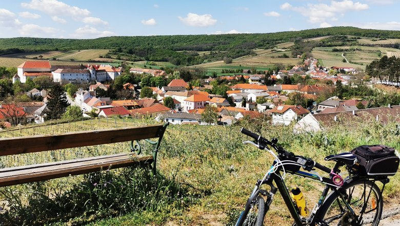Fahrrad und Bank mit Blick auf ein Dorf in hügeliger Landschaft.