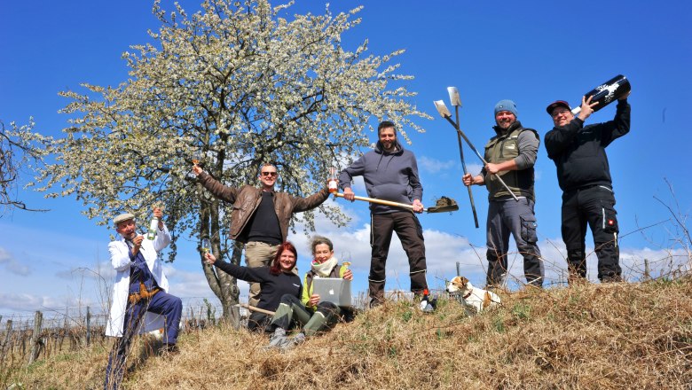 Gruppe von Menschen mit Werkzeugen und Getränken vor einem blühenden Baum im Freien.
