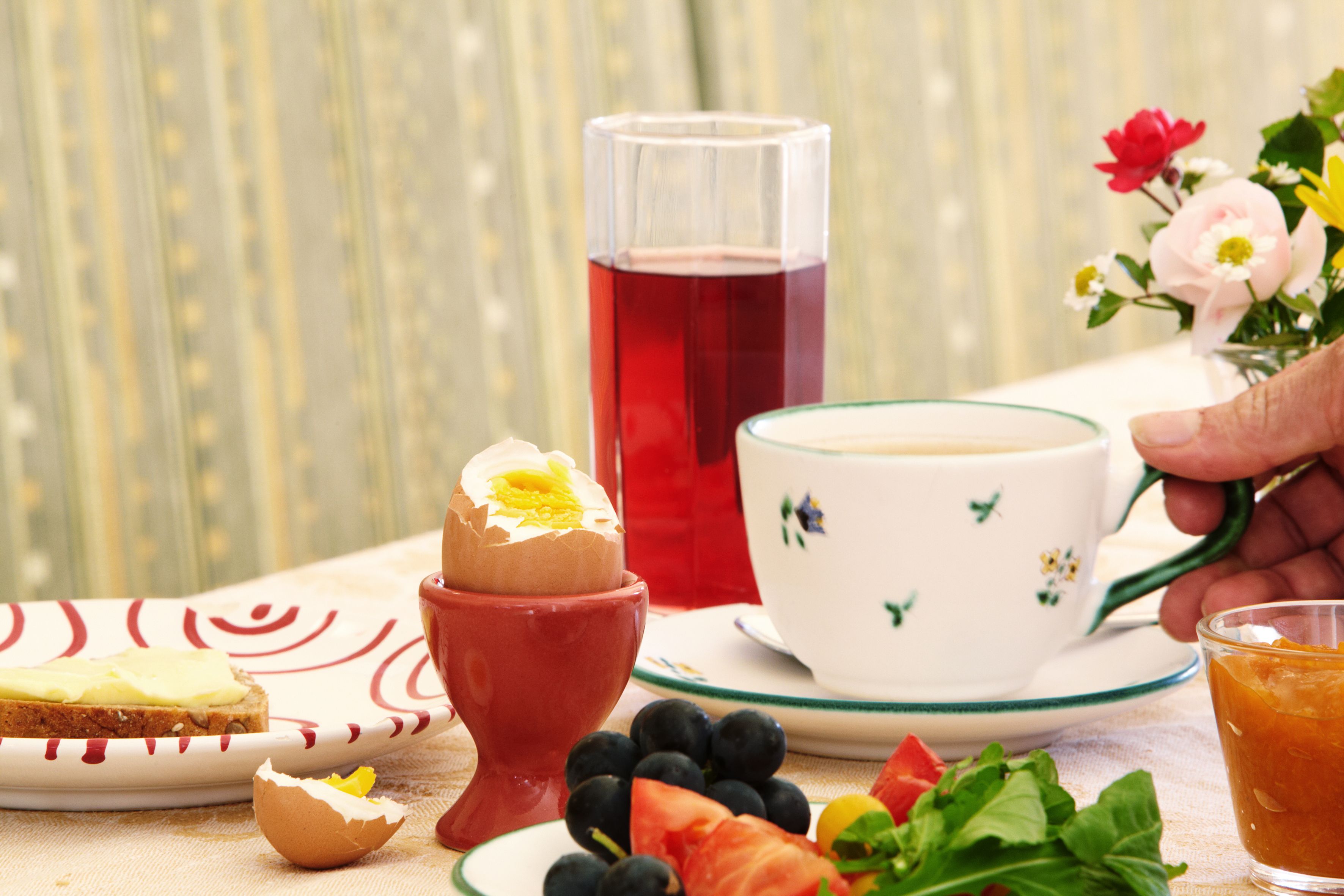 Frühstückstisch mit gekochtem Ei, Saft, Kaffee, Brot, Obst und Blumen.