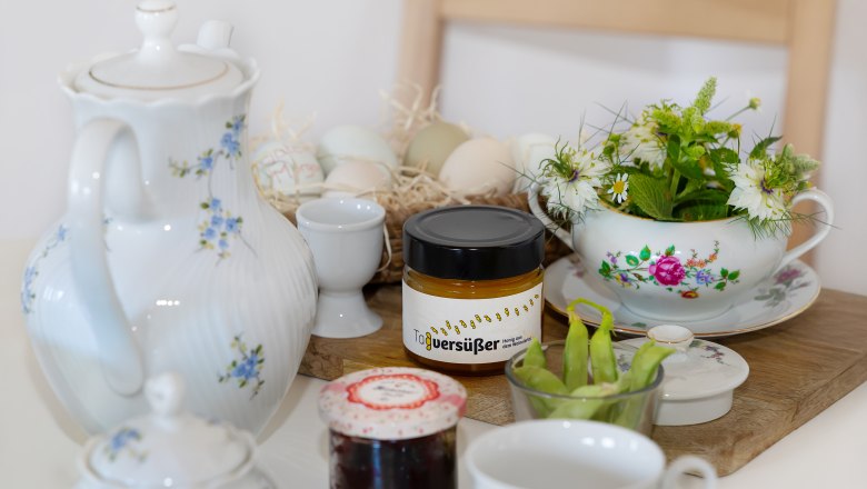 Frühstückstisch mit Teekanne, Honigglas, Marmelade, Eiern und Blumen.