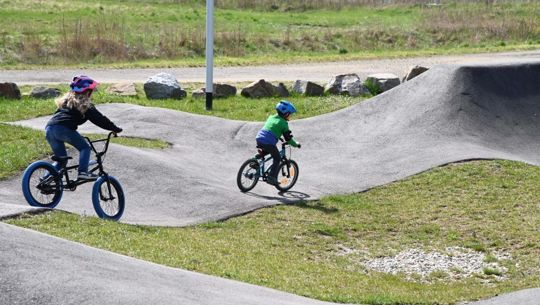 Zwei Kinder fahren mit Fahrr&auml;dern auf einem Pumptrack im Freien.