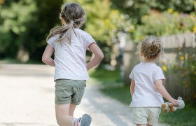 Zwei Kinder laufen auf einem Kiesweg in einem Garten, umgeben von Bäumen und Blumen.