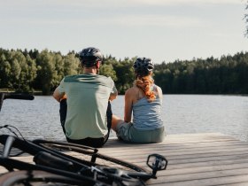 Zwei Radfahrer:innen mit Helmen sitzen an einem Steg und blicken in die Ferne, hinter ihnen die Fahrr&auml;der.