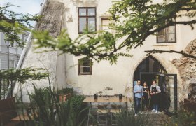 Drei Personen stehen in einem historischen Innenhof vor einem Gebäude mit gotischem Bogenfenster.