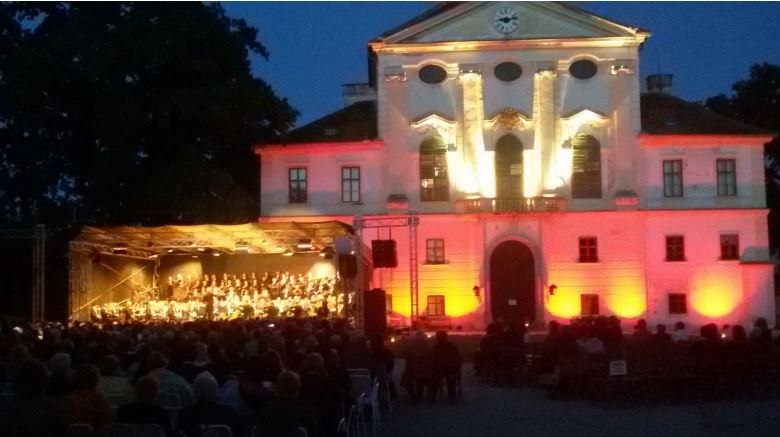 Ein beleuchtetes Geb&auml;ude mit einem Orchester, das auf einer B&uuml;hne im Freien spielt, umgeben von einem sitzenden Publikum bei Nacht.