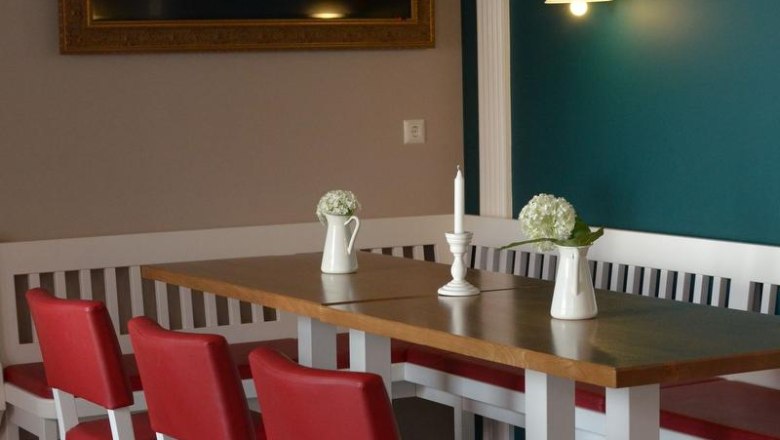 Ein Essbereich mit roten Stühlen, einem Holztisch, weißen Vasen und einer Kerze. An der Wand ein großer, leerer Bilderrahmen und eine Lampe.