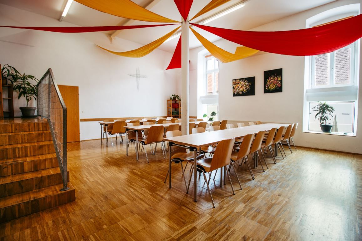 Svetlá jedáleň s drevenou podlahou, stolmi a stoličkami. Zo stropu visia červené a žlté textilné panely.