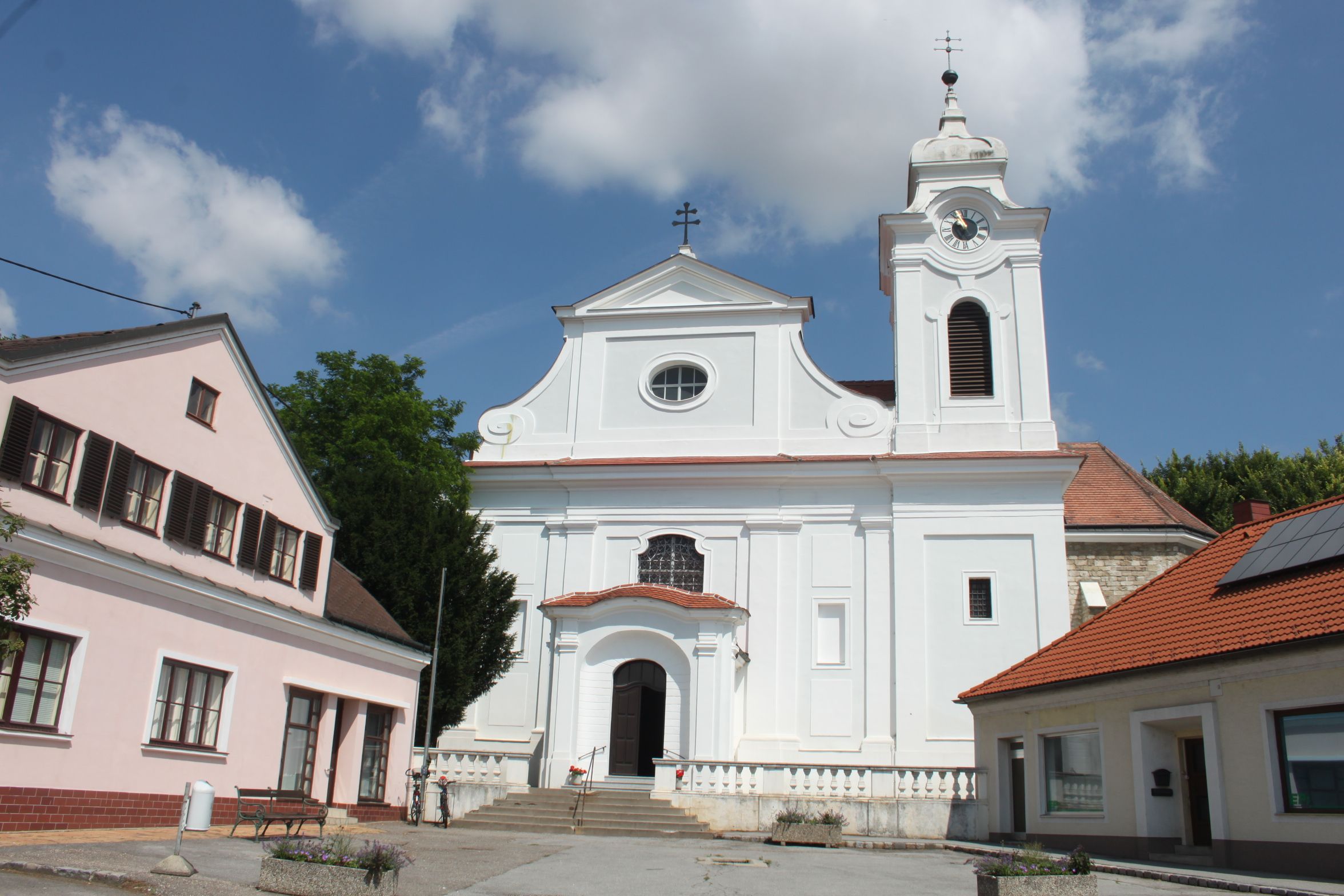 Farní kostel ve Wilfersdorfu s okolními budovami za slunečného počasí.
