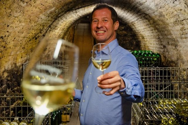 Ein Winzer mit einem Glas Weißwein in der Hand prostet in Richtung Betrachter:in.