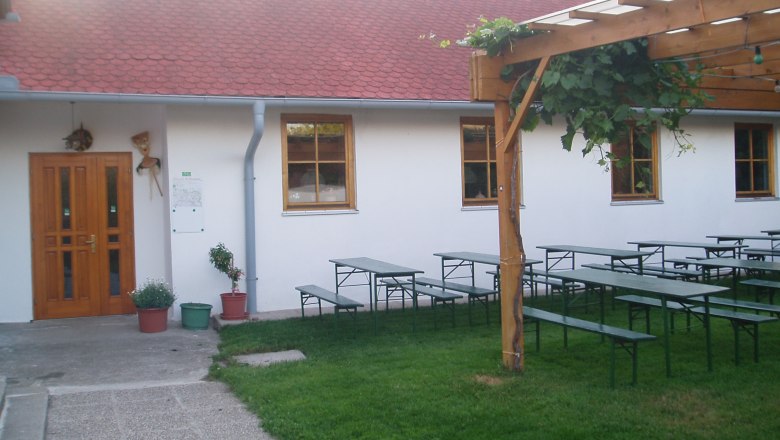 Ein Garten mit Biertischen, einem Holzpergola und einem Geb&auml;ude mit roten Dachziegeln.