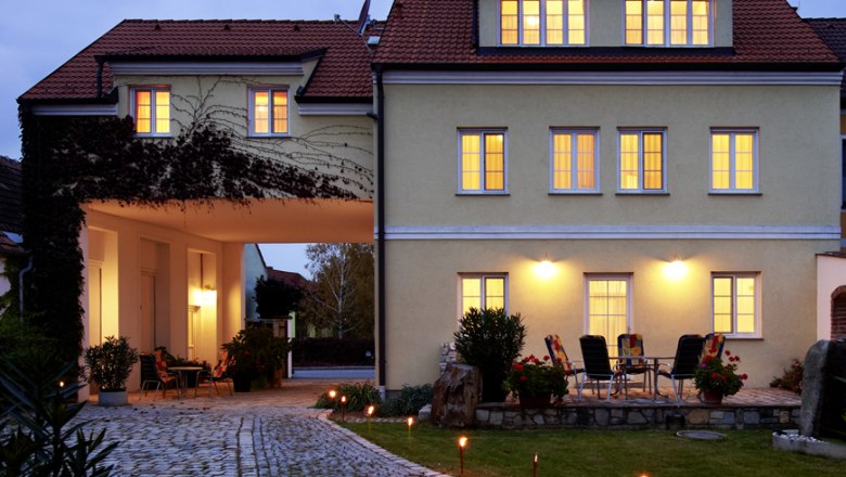 Beleuchtetes Gebäude mit gepflastertem Weg und Gartenmöbeln im Vordergrund.