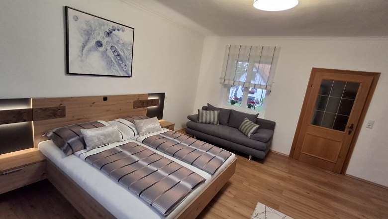 Modernes Schlafzimmer mit Doppelbett, grauem Sofa, Holzfußboden und abstraktem Kunstwerk an der Wand.
