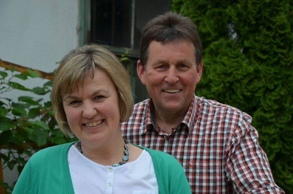 A smiling couple stands outside in front of a green hedge.