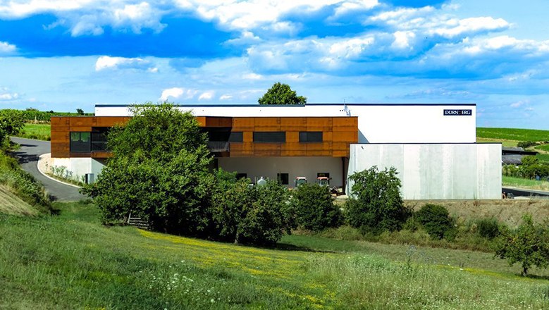 Moderne Weingut-Gebäude mit brauner und weißer Fassade, umgeben von grüner Landschaft und blauem Himmel.