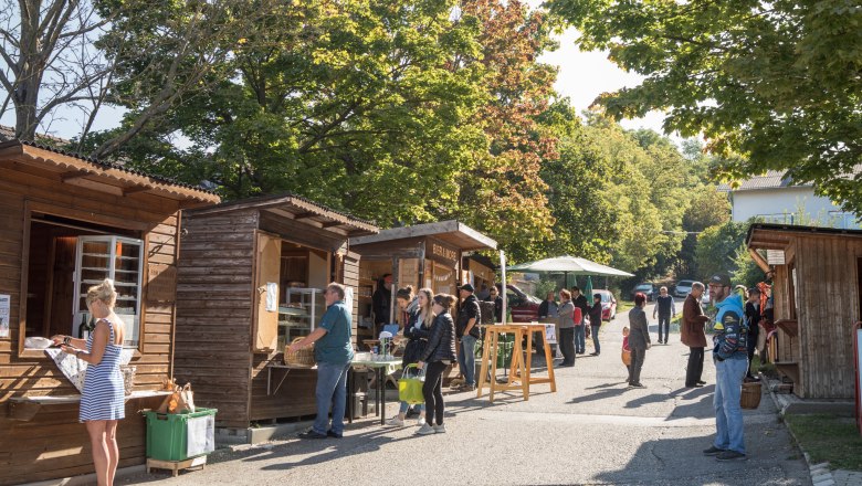 Menschen stehen an Holzhütten auf einem Markt im Freien bei sonnigem Wetter.