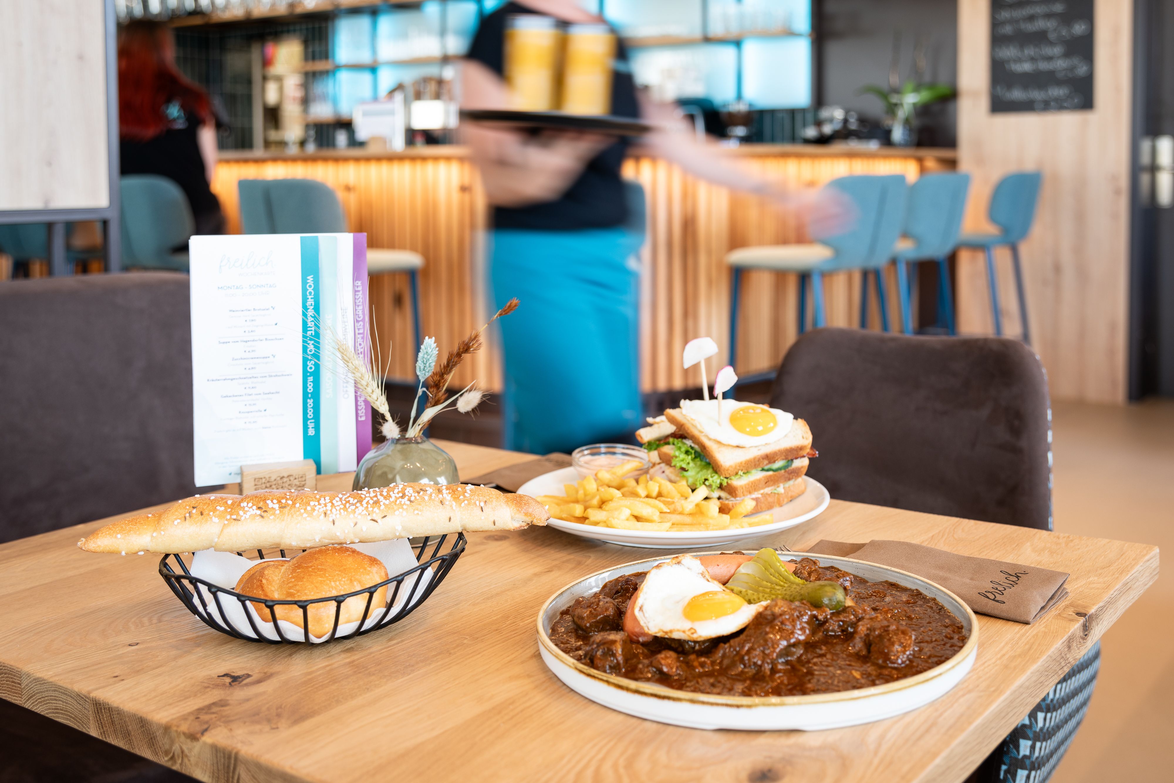 Ein Tisch in einem Restaurant mit regionalen Speisen: ein Teller mit Gulasch und Spiegelei, ein Sandwich mit Pommes und ein Brotkorb.