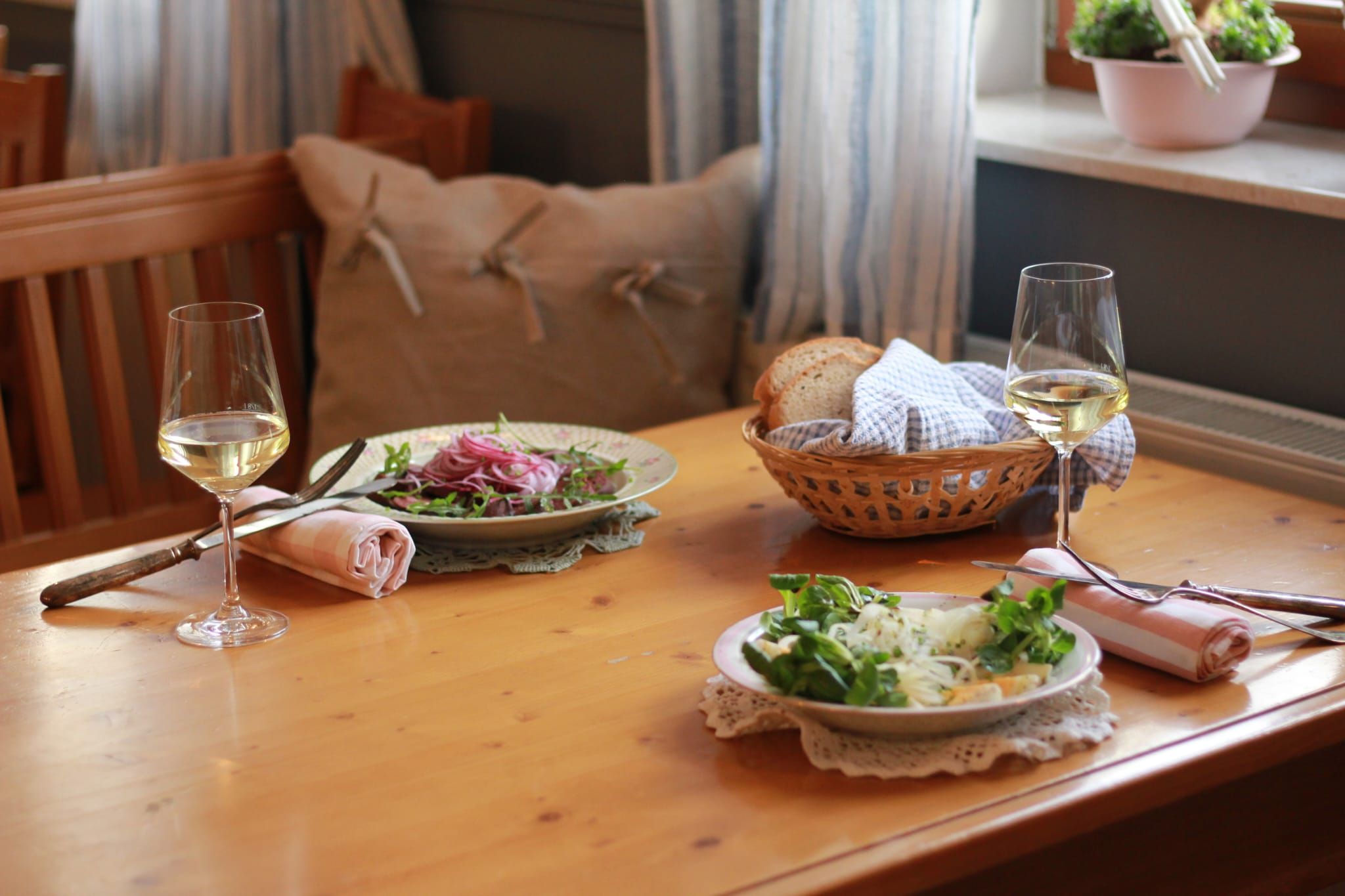 Zwei Teller mit Salat und zwei Gläser Weißwein auf einem Holztisch, daneben ein Brotkorb.