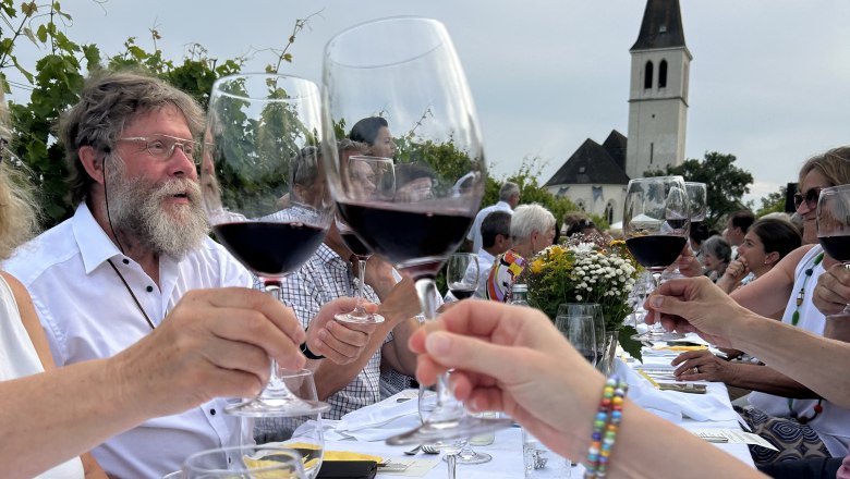 Menschen stoßen mit Weingläsern an einem langen Tisch im Freien an, im Hintergrund eine Kirche.