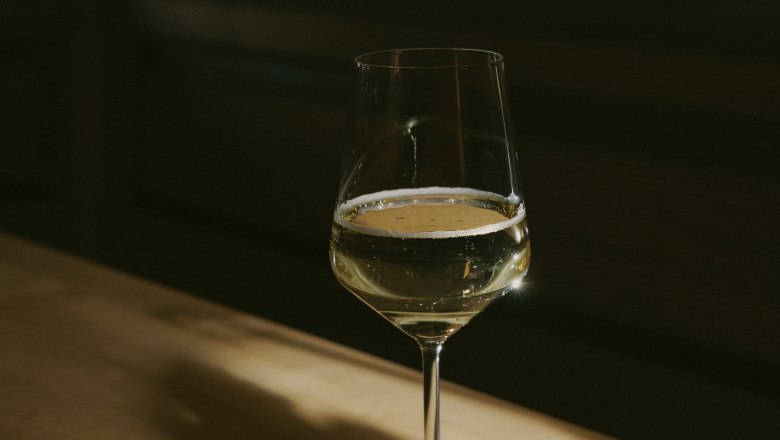 A wine glass with white wine stands on a wooden table in the shade.