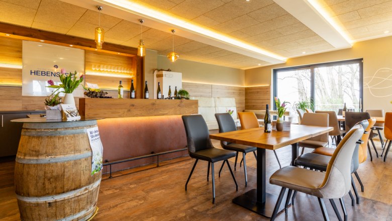 Moderner Verkostungsraum mit Holztischen, St&uuml;hlen und einer Bar. Weinflaschen und Blumen dekorieren den Raum.