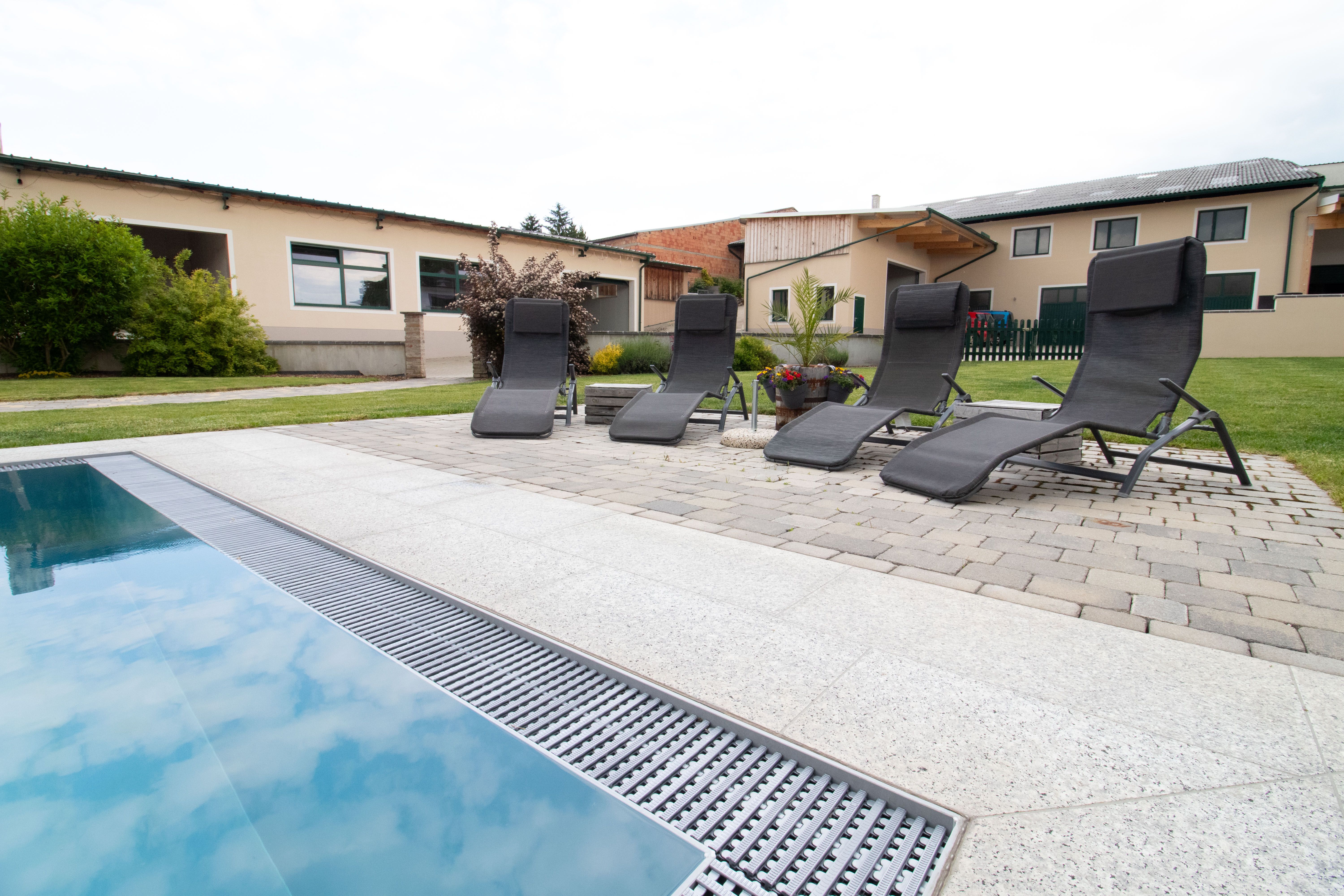 Vier Liegestühle stehen neben einem Pool auf einer Terrasse mit Pflastersteinen. Im Hintergrund sind Gebäude und ein Garten zu sehen.