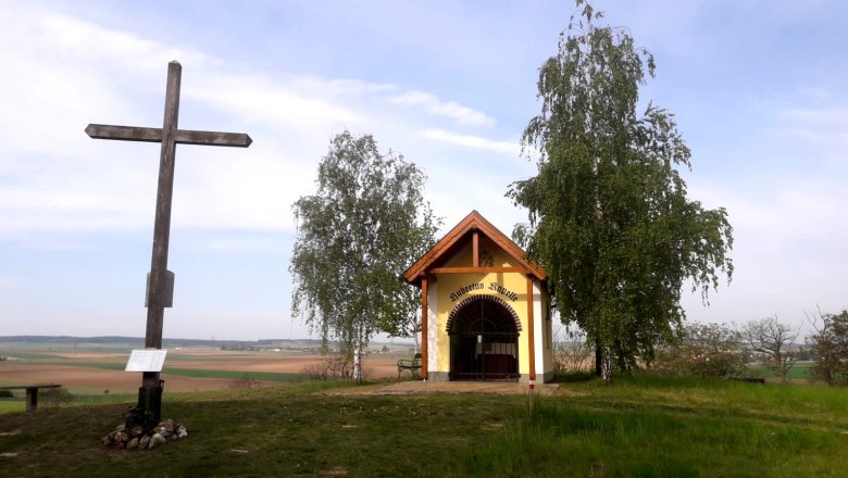 Ein Holzkreuz und eine kleine Kapelle auf einem H&uuml;gel mit B&auml;umen und weitem Blick &uuml;ber die Landschaft.