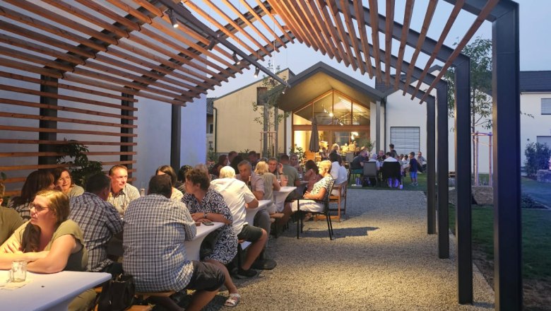 Menschen sitzen an langen Tischen unter einem Holzpergola bei Abenddämmerung.