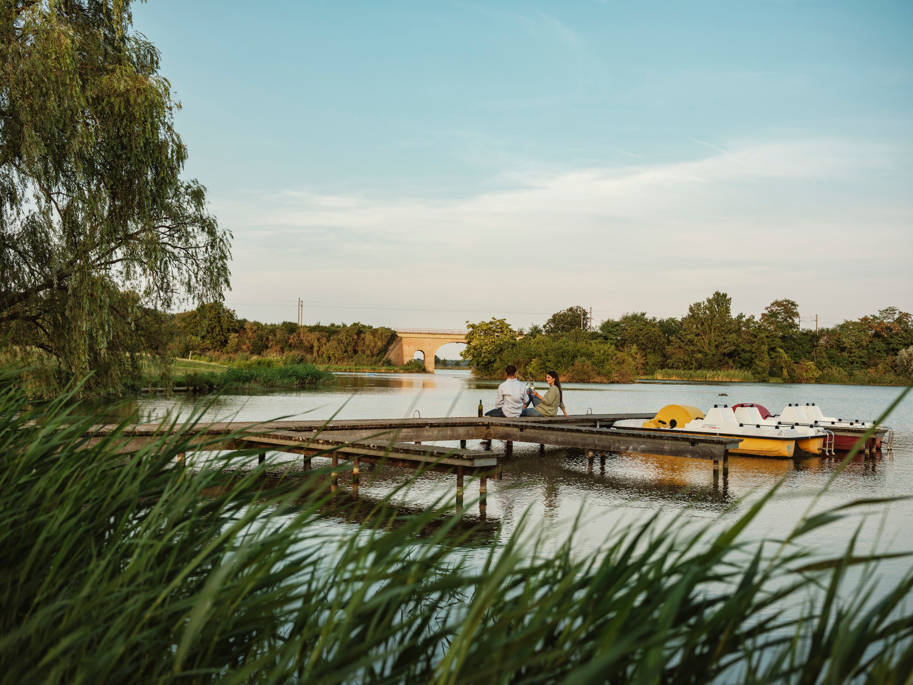 Ein Paar sitzt auf einem Steg am Teich in Bernhardsthal, umgeben von Natur und Tretbooten.