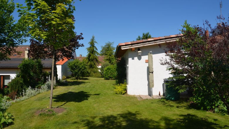 Ein gepflegter Garten mit Rasen, Bäumen und Sträuchern. Im Hintergrund sind zwei kleine Gebäude mit roten Dächern zu sehen.