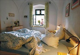 Gem&uuml;tliches Schlafzimmer mit ungemachtem Bett und Blick aus dem Fenster auf eine Person mit Blumen.