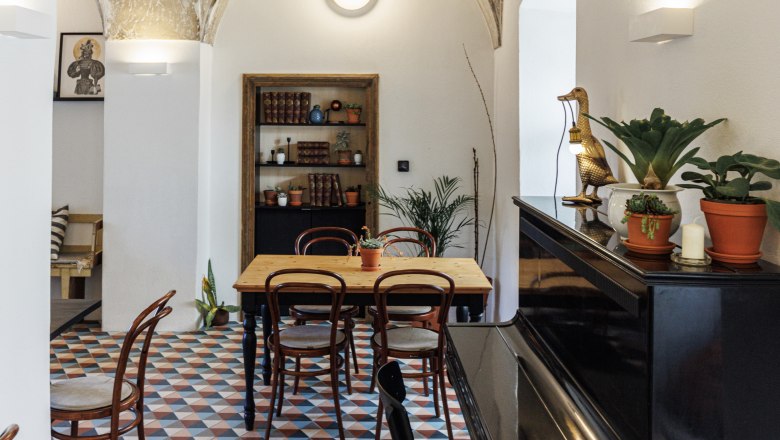 Ein stilvolles Café mit gewölbter Decke, buntem Fliesenboden, Holztisch und Stühlen. Ein Klavier steht rechts, Pflanzen und Dekorationen sind verteilt.