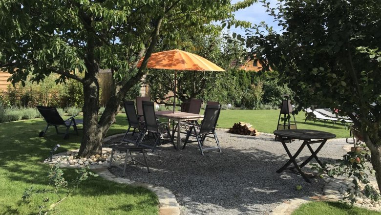Garten mit Sitzbereich unter Bäumen, Tisch und Stühlen, orangefarbener Sonnenschirm.